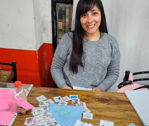 Una persona sentada en una mesa de madera con varias tarjetas ilustradas, una hoja azul con el texto 'Tarjetas Rutinas', un teléfono blanco y otros pequeños objetos.