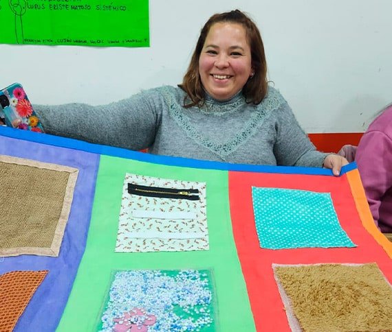 Persona sosteniendo una colcha colorida compuesta por seis cuadrados con texturas y patrones diferentes, frente a un cartel informativo sobre el Lupus Eritematoso Sistémico.