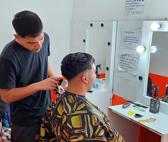 Chico joven practicando corte de pelo en el curso de barbería con otro chico sentado