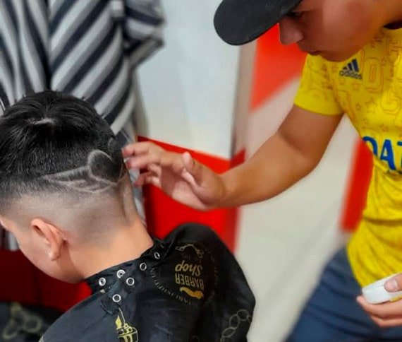 Chico joven practicando corte de pelo con dibujos en el curso de barbería con otro chico sentado