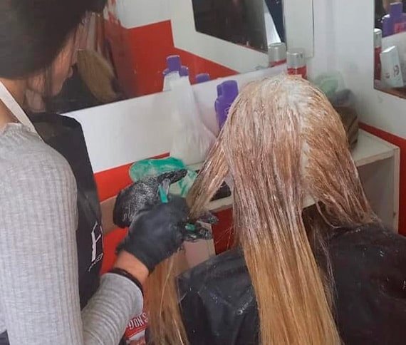 Persona aplicando un tratamiento para el cabello con guantes y brocha en un salón, con productos y espejo al fondo.
