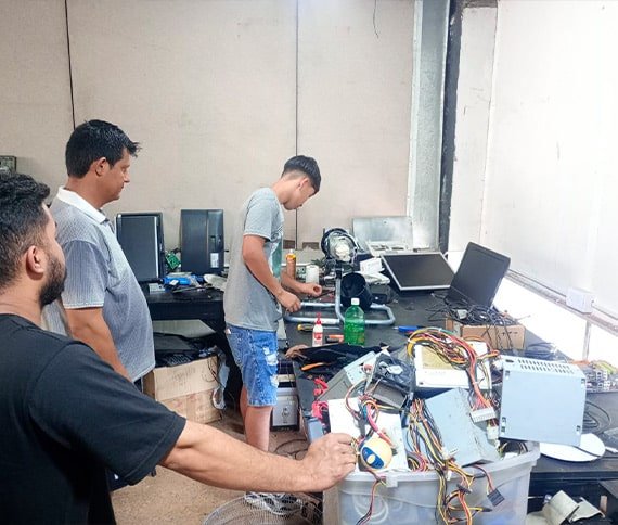 Tres personas trabajando alrededor de una mesa, llena de componentes electrónicos y herramientas. La mesa tiene varios monitores de computadora, una laptop, una botella de pegamento y cables. Parece ser un entorno de taller técnico o de reparación, iluminado con luz natural que entra por una ventana grande.