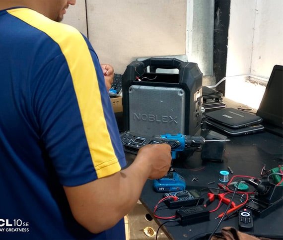 Una persona trabajando en un puesto de trabajo. Sostiene un taladro inalámbrico azul, mientras parece estar reparando un dispositivo electrónico. En la mesa hay varias herramientas y componentes electrónicos, como un soldador, cables, un multímetro y un rollo de estaño. Junto con algunas laptops y otros dispositivos electrónicos. La imagen muestra un proceso de reparación o ensamblaje de equipo electrónico.