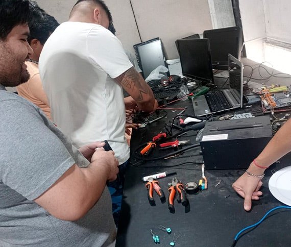 Un grupo de personas trabajando en una mesa llena de herramientas electrónicas y componentes, como laptops, monitores de computadora, destornilladores, alicates, y circuitos. La escena sugiere un entorno técnico o de reparación relacionado con hardware o electrónica.