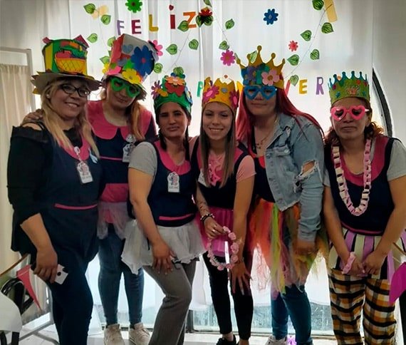 Grupo de seis personas con disfraces coloridos frente a un cartel que dice 'FELIZ PRIMAVERA', decorado con flores y hojas.