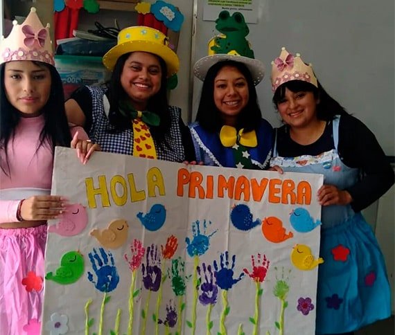 Grupo de cuatro personas sosteniendo un cartel que dice 'HOLA PRIMAVERA', con decoraciones coloridas que asemejan flores y pájaros.