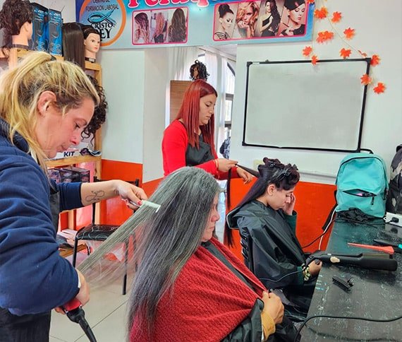 La imagen muestra dentro de un salón de belleza. Hay dos personas sentadas en sillas, cada una teniendo su cabello estilizado por dos diferentes estilistas. Una estilista usa una plancha para alisar el cabello de la persona a la izquierda, mientras que la otra estilista trabaja en el cabello de la persona a la derecha. La persona a la izquierda tiene el cabello largo y liso y está cubierta con una capa roja.