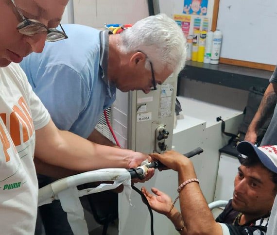 Tres personas trabajando juntas en una tarea de mantenimiento o reparación. Una de ellas sostiene una herramienta y trabaja en un equipo, mientras otra colabora sosteniendo una parte del mismo. La tercera observa o asiste desde un costado. El equipo parece estar relacionado con aire acondicionado o refrigeración, como indica la etiqueta 'R-410A' en el fondo. El escenario parece ser un taller, con varias herramientas y suministros visibles en estantes y una mesa de trabajo.