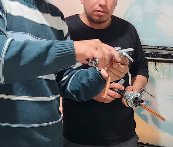 Dos personas trabajando con herramientas metálicas y tuberías de cobre, enfocándose en sus manos y las herramientas.