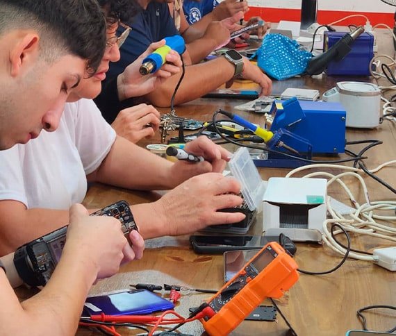 Grupo de personas reparando dispositivos electrónicos sobre una mesa llena de herramientas como soldadores, multímetros y componentes electrónicos.