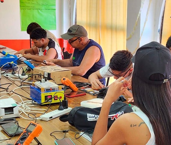 Personas trabajando en una mesa con herramientas electrónicas como soldadores, cables y componentes, en un ambiente de taller o aula.