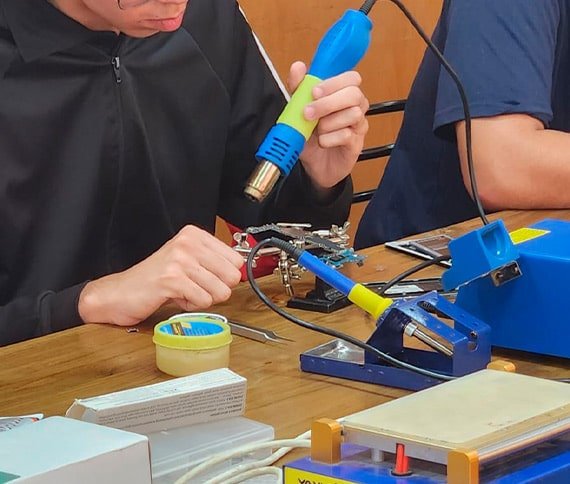 Personas trabajando en un proyecto de electrónica, utilizando herramientas como pistola de calor, soporte para circuitos, soldador y cinta adhesiva en una mesa de trabajo.