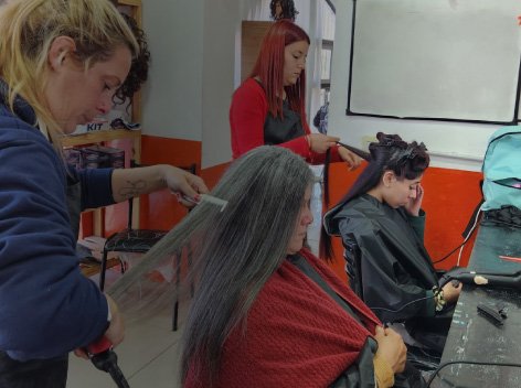 Estudiantes de peluquería practicando técnicas de peinado y alisado en un salón de clases equipado con espejos y herramientas de estilismo.