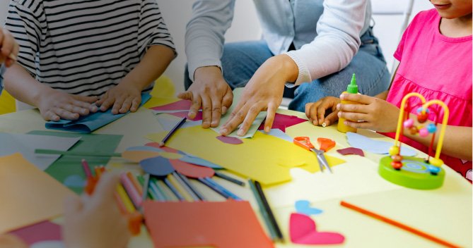 Niños y adultos participando en una actividad de manualidades con papel de colores, tijeras, pegamento y lápices sobre una mesa.