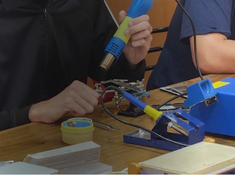 Personas realizando trabajos de soldadura electrónica en un taller, utilizando una estación de soldadura y una pistola de calor.
