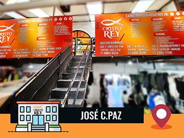 Imagen de la sucursal de José C. Paz, un espacio interior con dos grandes carteles naranjas con texto blanco. Los carteles corresponden a 'Cristo Rey' y detallan servicios y cursos ofrecidos, como computación, peluquería, cocina, mecánica, entre otros. En la parte inferior de la imagen hay un logo de 'Cristo Rey' y texto que dice 'José C. Paz' acompañado de un ícono de ubicación.