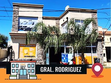 Edificio moderno de dos pisos de la Sucursal en Gral. Rodríguez con detalles de piedra y amplias ventanas. En la parte frontal hay palmeras que adornan el paisaje. A la izquierda del edificio, se encuentra un gran anuncio publicitario que muestra los servicios ofrecidos.